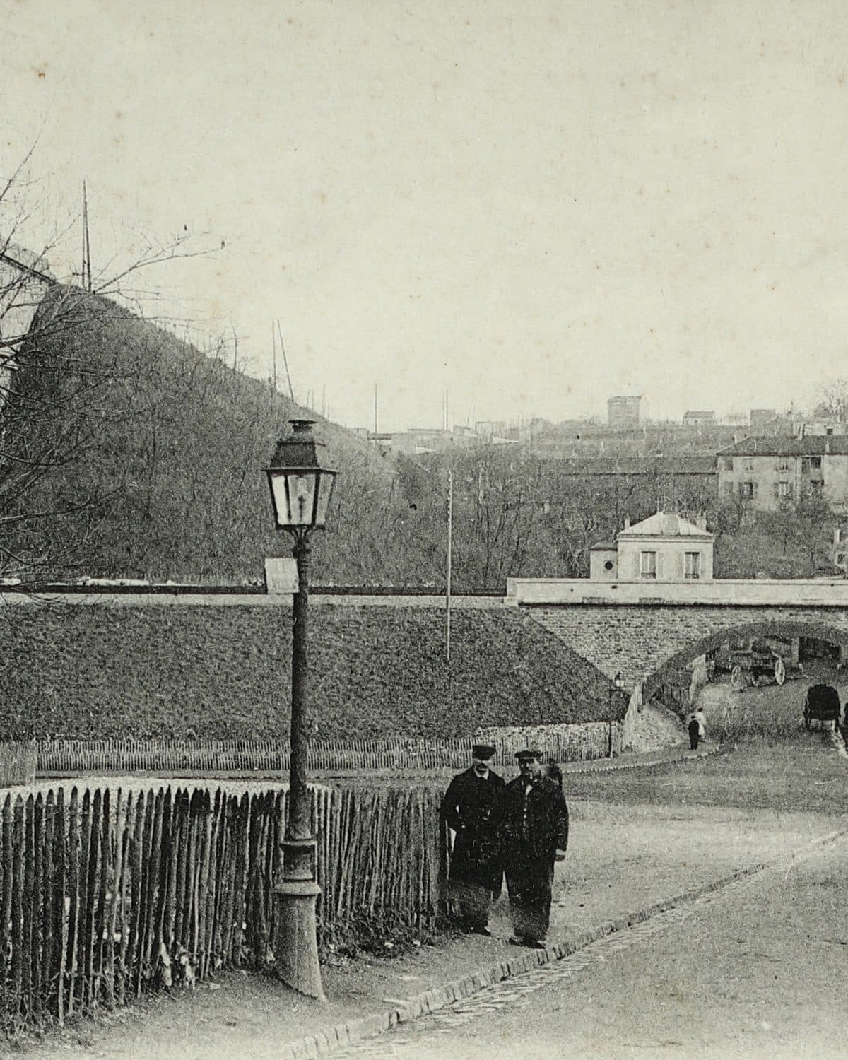 Carte postale vers 1900 : la rue descend vers le Val Fleury sous un pont, au pied du talus du viaduc ; deux passants près d'un réverbère.